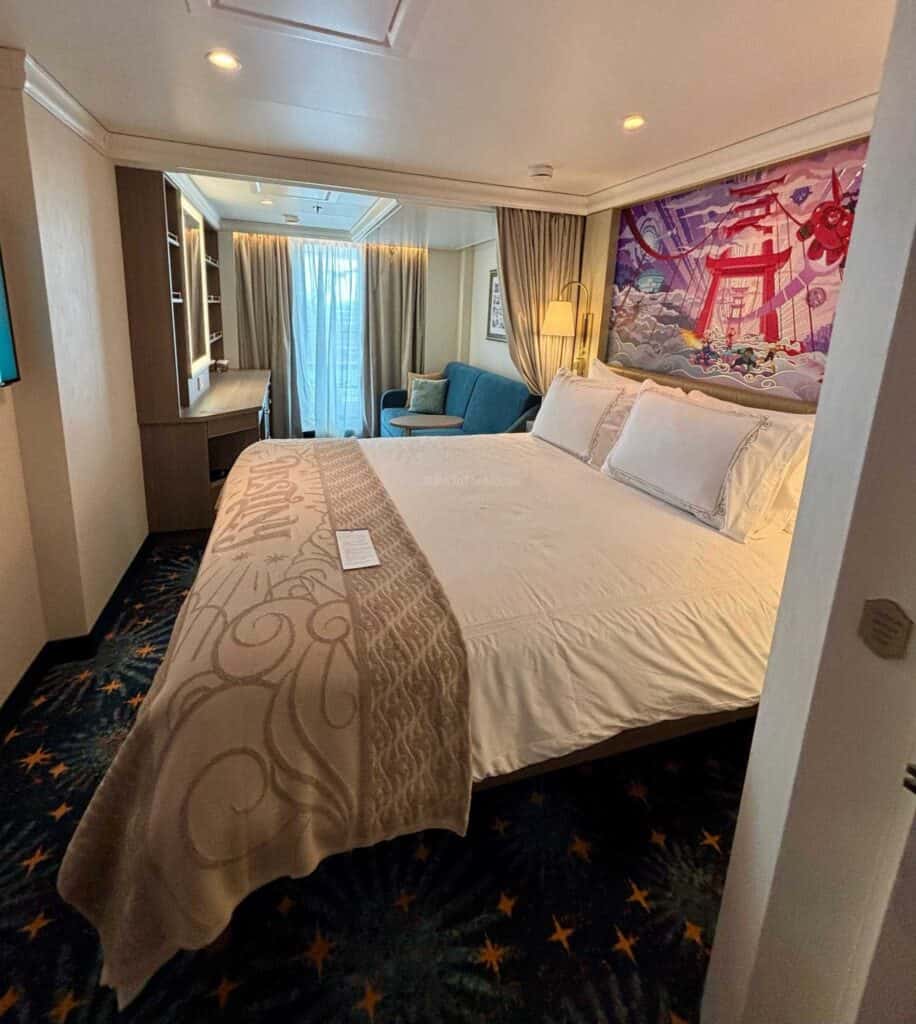 Interior view of a deluxe family oceanview stateroom on the Disney Destiny, featuring a queen-sized bed with decorative bedding, a small seating area with a blue sofa, and large windows with sheer curtains.