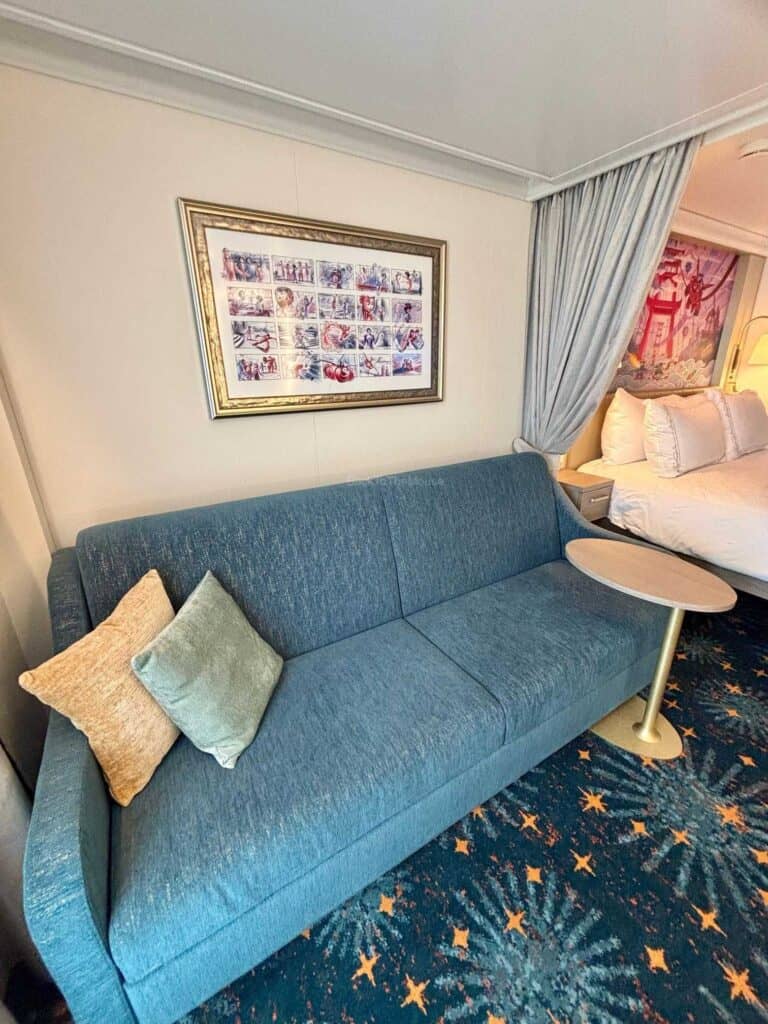 Interior view of a Disney cruise ship stateroom featuring a blue sofa with pillows, a small round table, and a framed artwork on the wall.