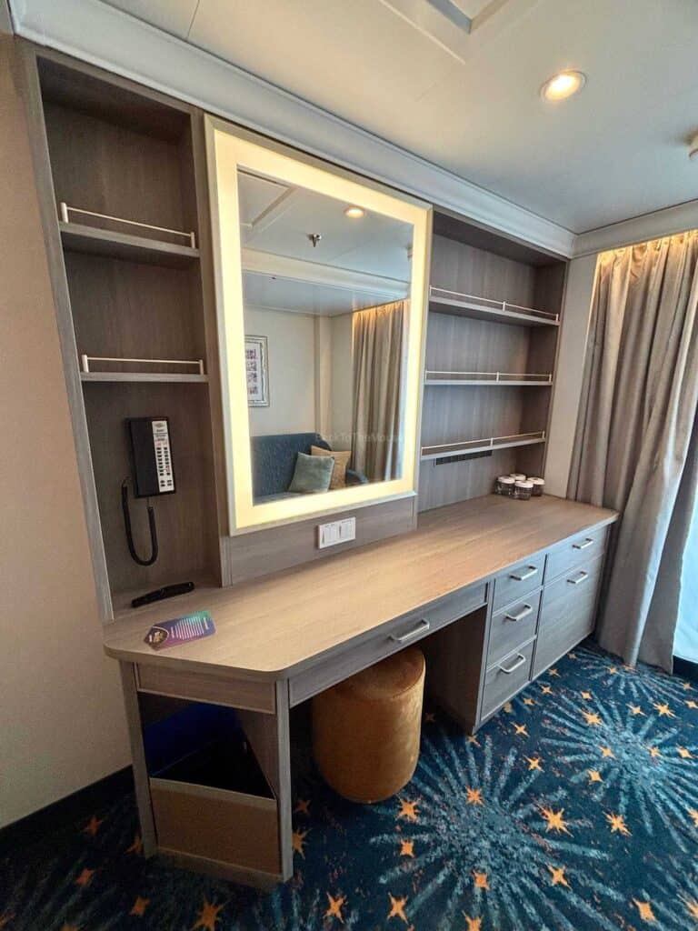 Interior view of a stateroom on Disney Destiny showing a wooden desk with a mirror, shelves, and decorative carpet.