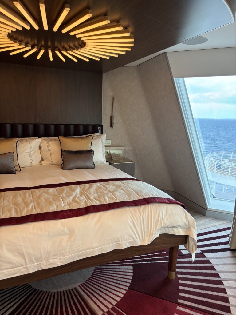 A luxurious stateroom featuring a king-sized bed with elegant linens and decorative pillows, a modern overhead light fixture, and a large panoramic window with ocean views.