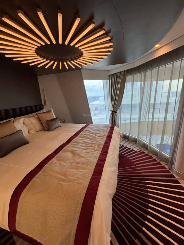 Interior view of a luxury stateroom on the Disney Destiny cruise ship, featuring a large bed with decorative cushions, an intricate ceiling light, and large windows with curtains.