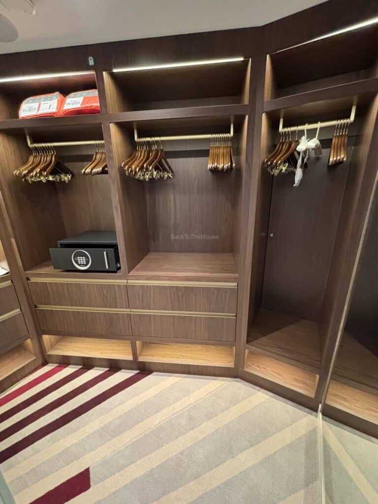 Interior view of a cruise ship stateroom closet featuring wooden hangers, shelves, a safe, and a patterned carpet.