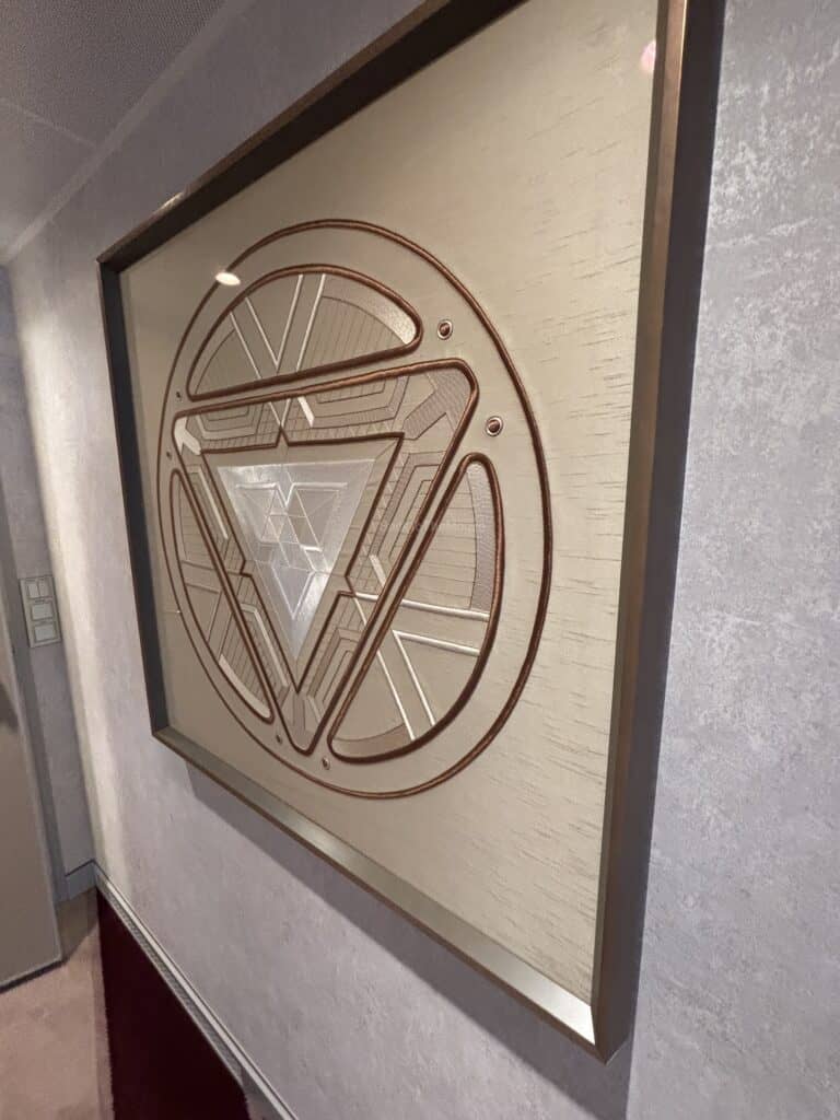 A framed decorative wall art piece featuring a geometric design resembling a logo, displayed in the hallway of a cruise ship suite.