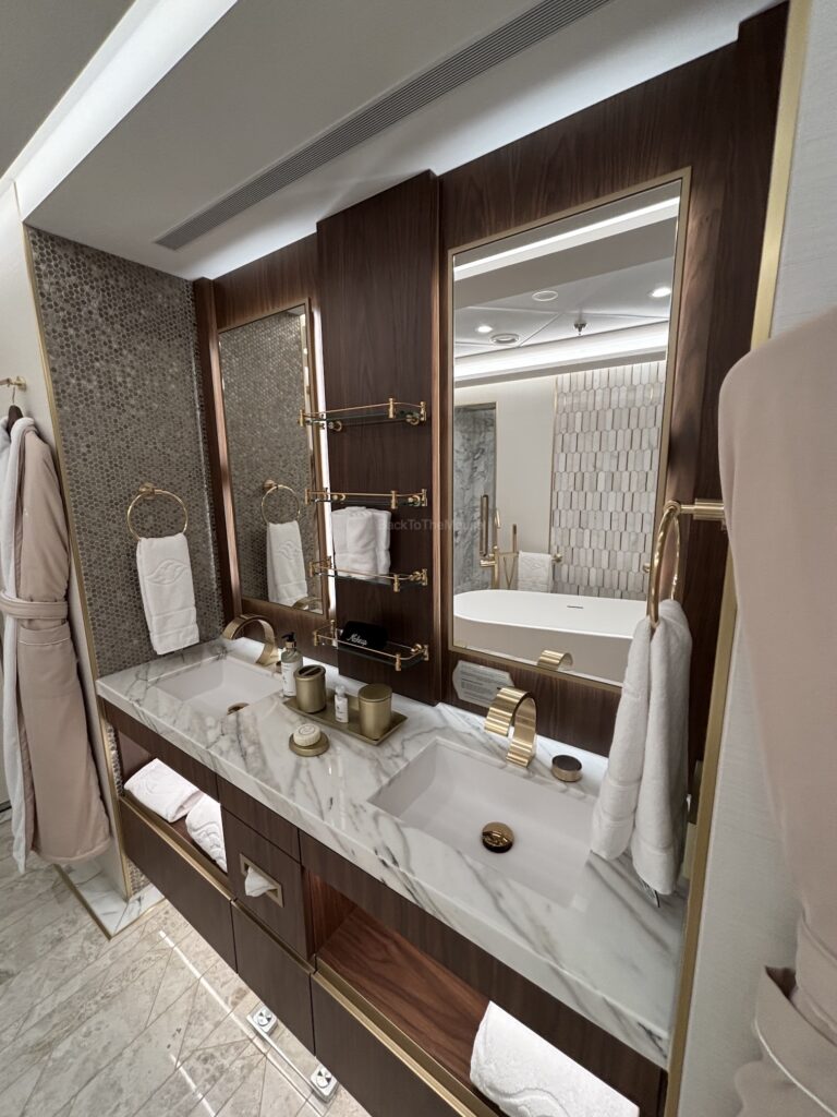 A luxurious bathroom with a marble double sink, gold fixtures, and elegant decor, featuring a large mirror and various toiletries.