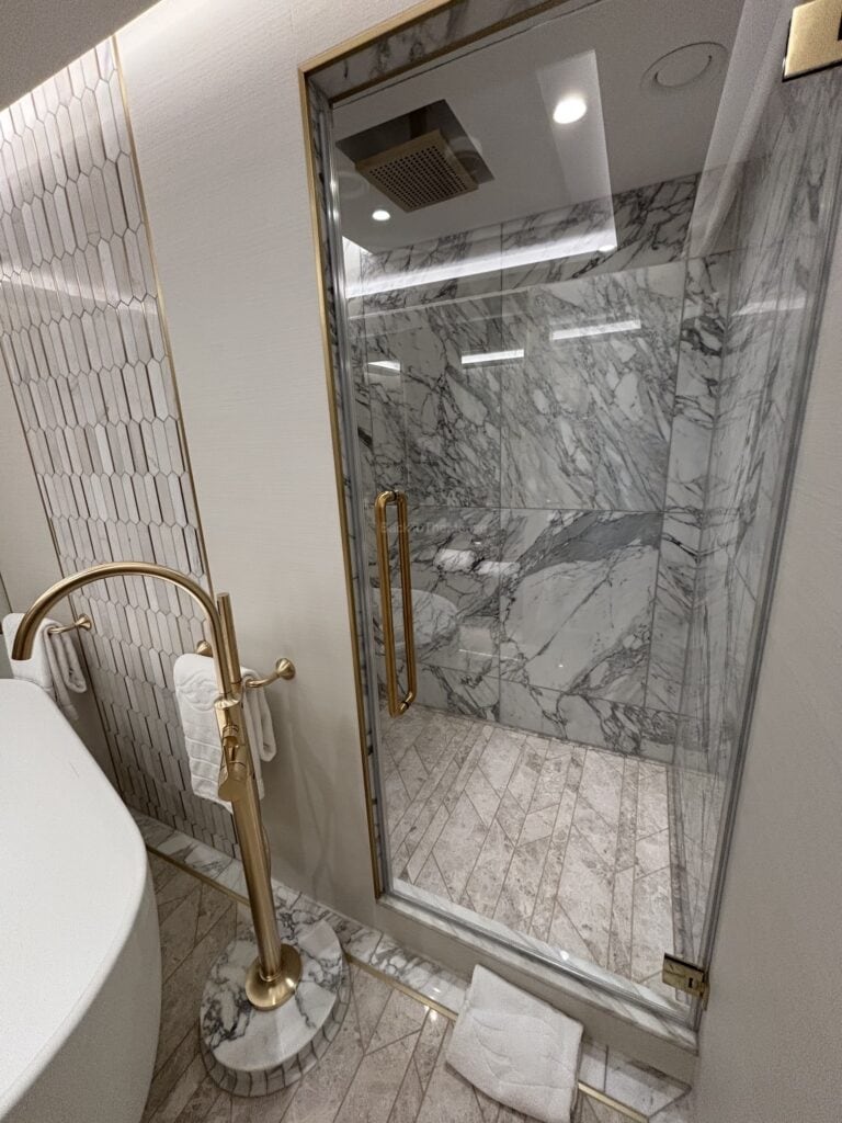 A luxurious bathroom featuring a glass shower enclosure with marble walls, a freestanding bathtub, and gold fixtures.