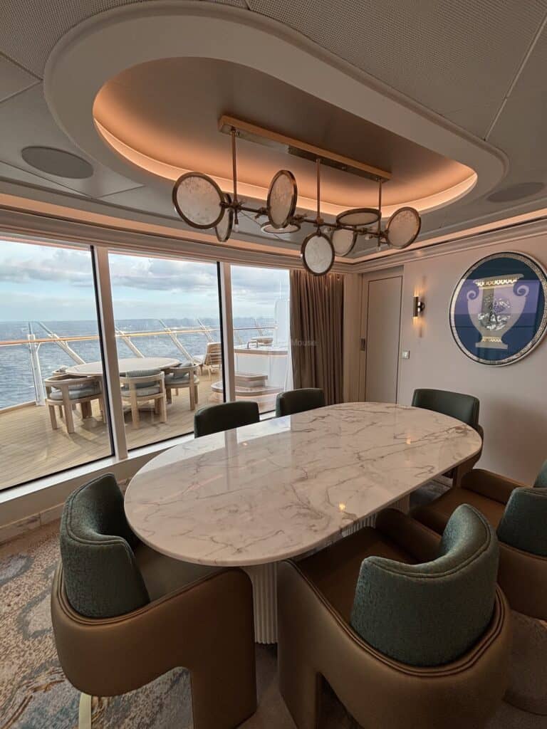 Interior view of a luxurious dining area in a cruise ship suite, featuring a round marble table surrounded by stylish chairs and large windows offering a view of the sea.