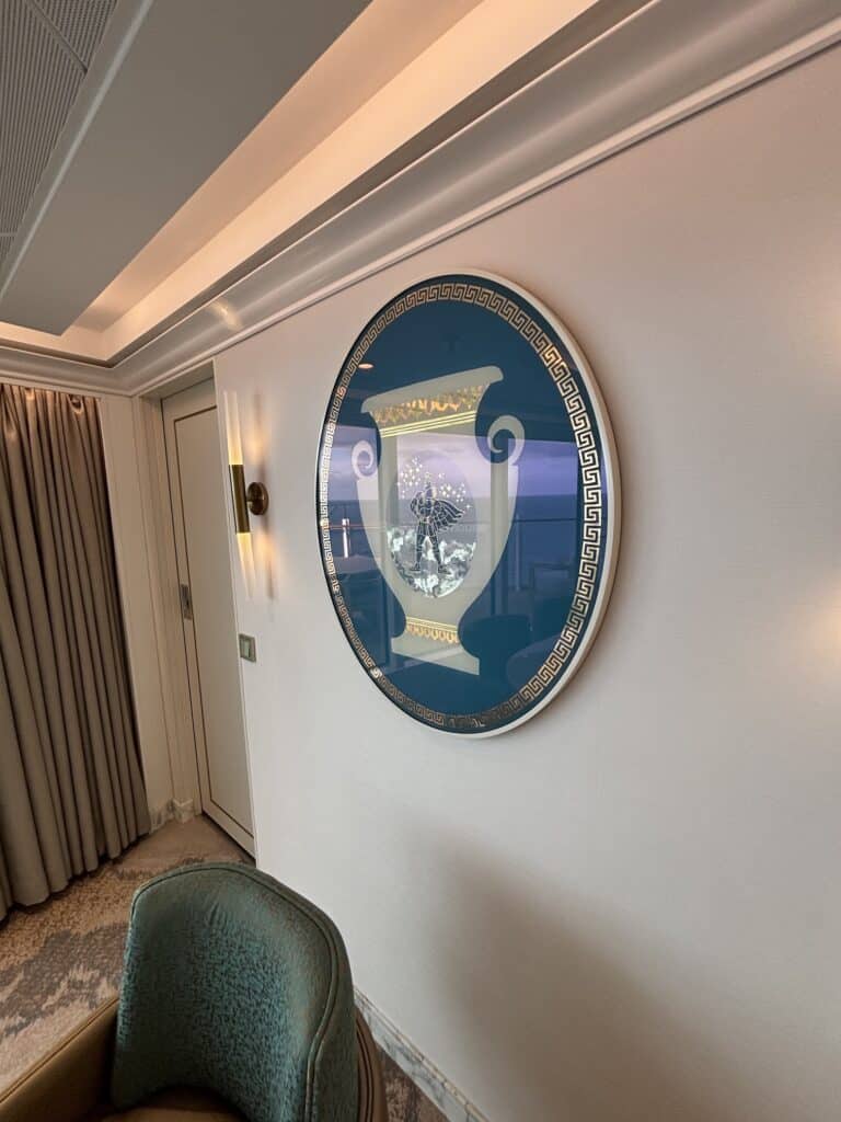 A decorative round wall art piece featuring a stylized vase design, located on the wall of a cruise ship stateroom, with soft lighting and curtains in the background.