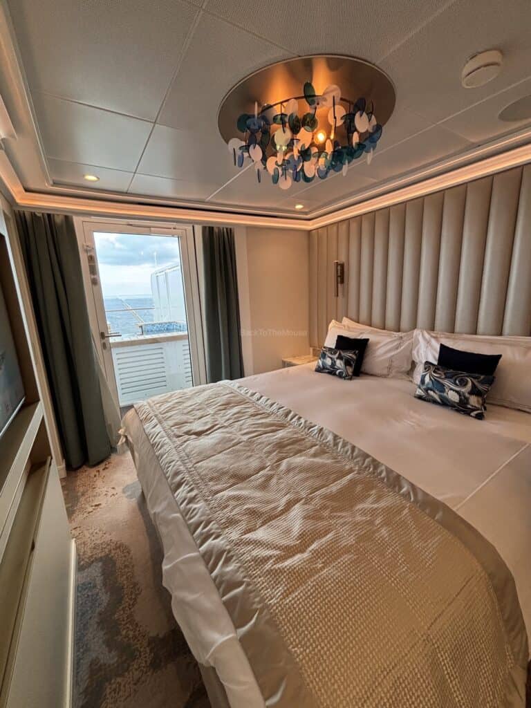 Interior view of a Disney cruise stateroom featuring a large bed with decorative pillows, modern light fixture, and a window with ocean view.