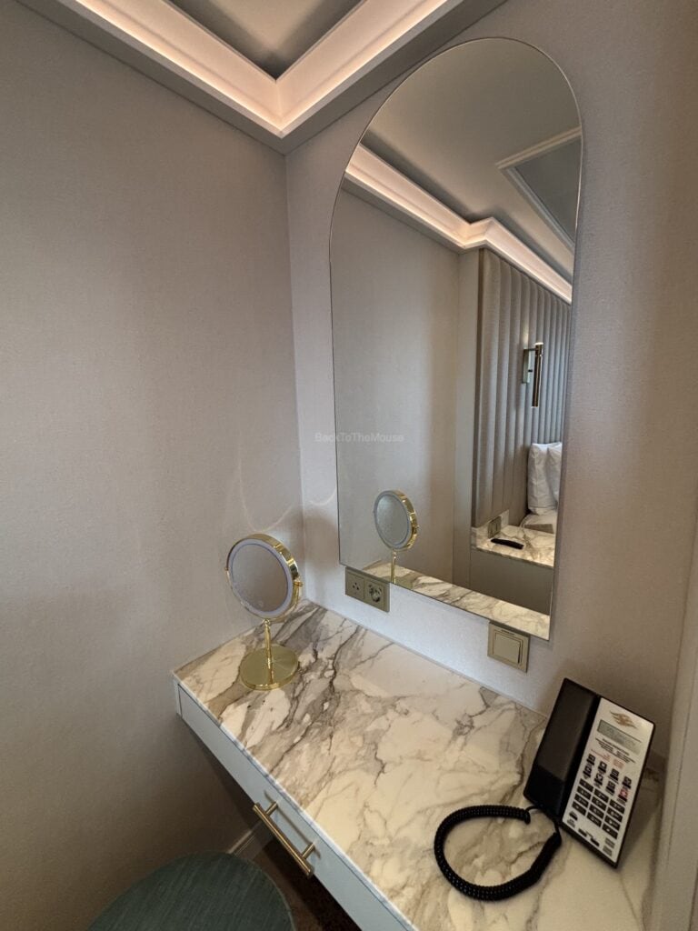 Elegant vanity area in a stateroom featuring a marble countertop, a round mirror with light, a small magnifying mirror, and a phone.
