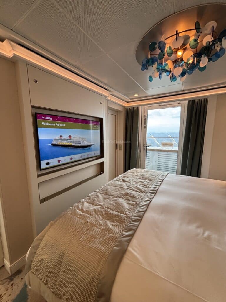 Interior view of a stateroom on the Disney Destiny cruise ship, featuring a large bed, modern decor, wall-mounted television displaying 'Welcome Aboard,' and a window with ocean view.
