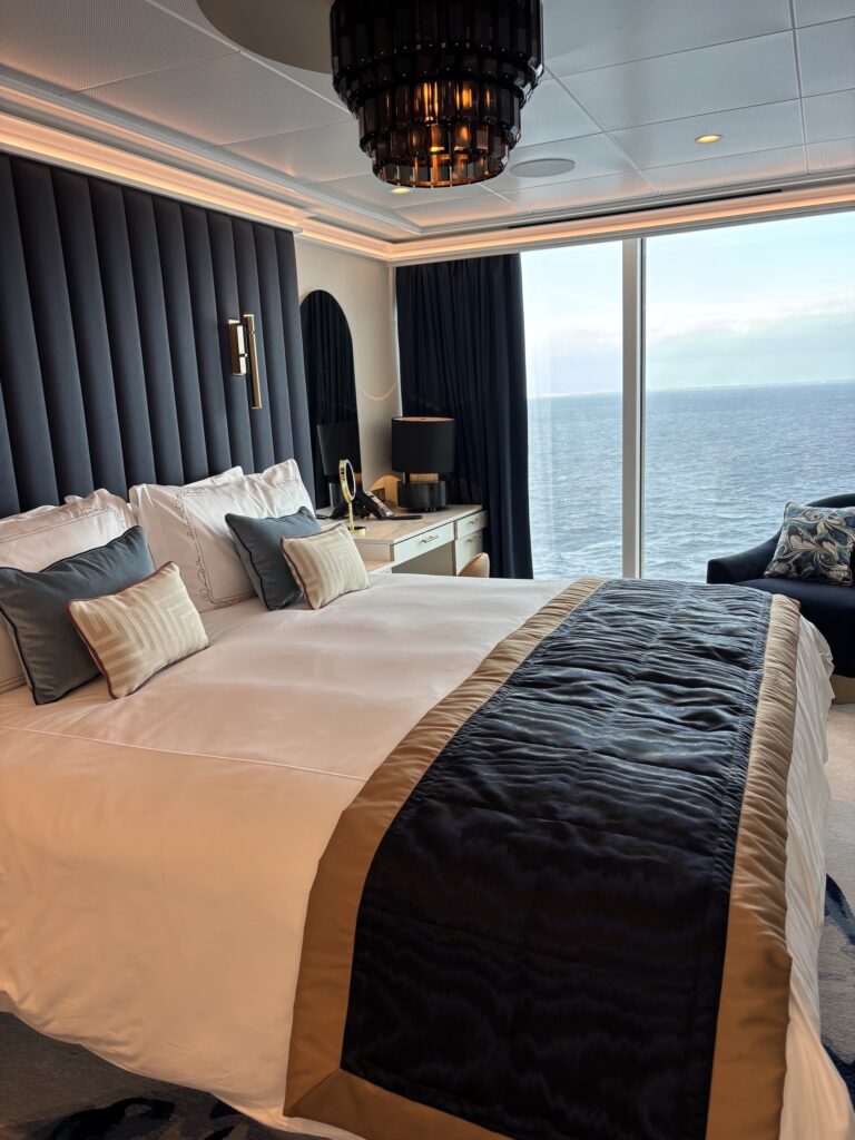 A luxurious cruise stateroom featuring a large bed with decorative pillows, elegant lighting, and a window showcasing the ocean view.