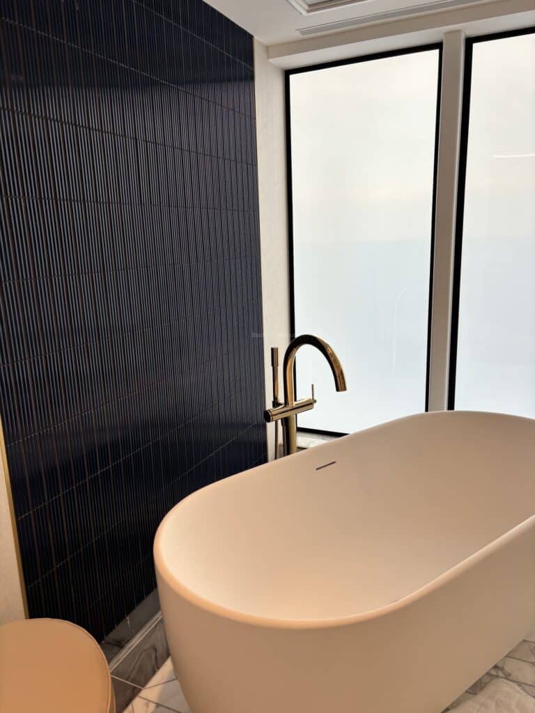 A modern bathroom featuring a freestanding bathtub beside a large window with a view. The wall behind the tub is covered in dark blue tiles, and there is a stylish gold faucet.
