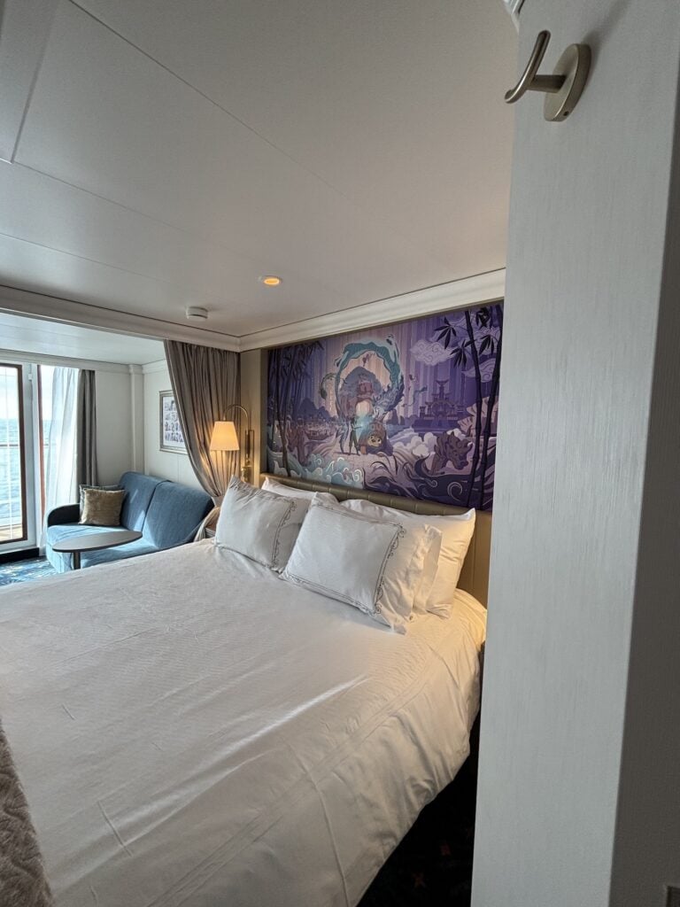 Interior view of a Deluxe Oceanview Stateroom with Verandah on the Disney Destiny, featuring a neatly made bed with white linens, a modern couch, and a decorative mural on the wall.