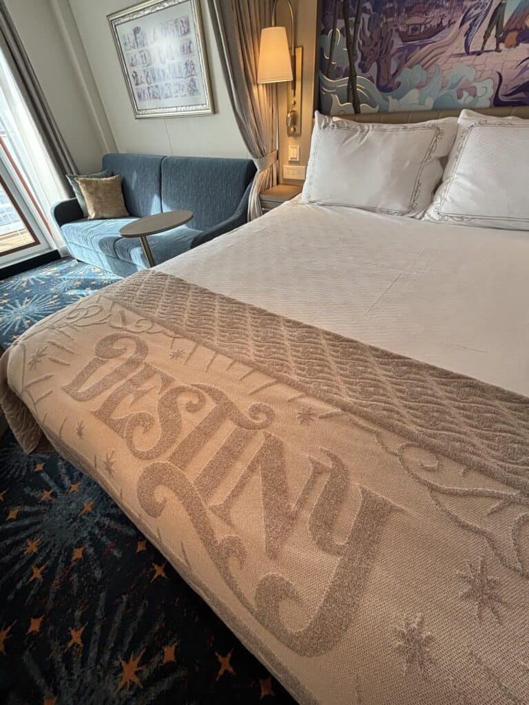 Interior view of a Disney cruise stateroom featuring a bed with a decorative blanket, a cozy blue sofa, and natural light from a large window.