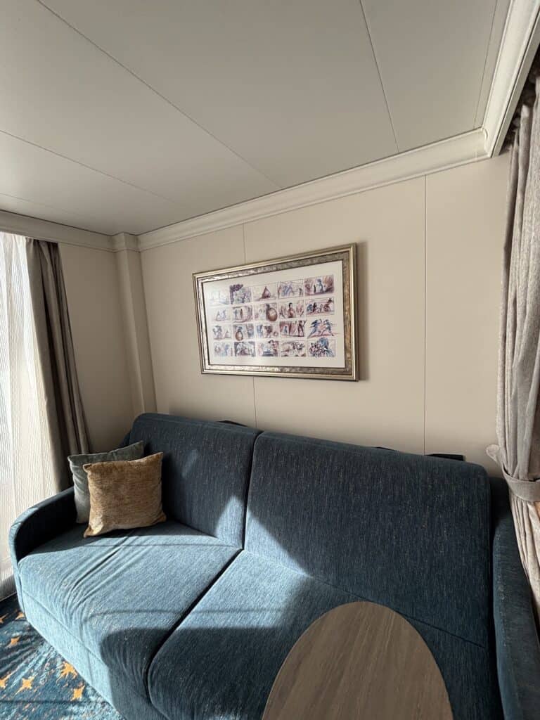 Interior view of a Deluxe Oceanview Stateroom with a blue couch, decorative pillows, and a framed picture on the wall. Natural light coming through the window.