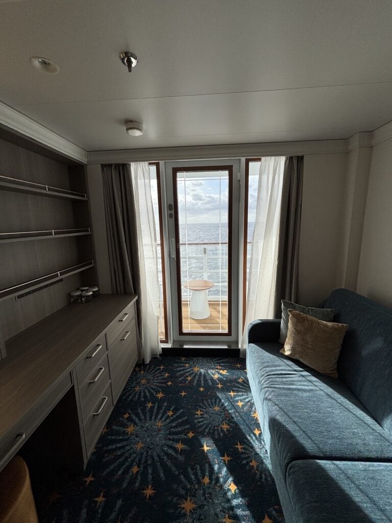 Interior view of a Deluxe Oceanview Stateroom on the Disney Destiny cruise ship, featuring a blue sofa, a desk with storage, and a large window with a view of the ocean.