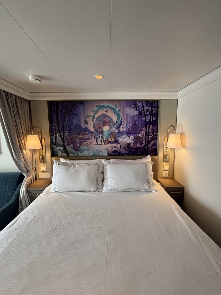 Interior of a Deluxe Oceanview Stateroom on the Disney Destiny, featuring a large bed, decorative wall art, and lamps on either side.