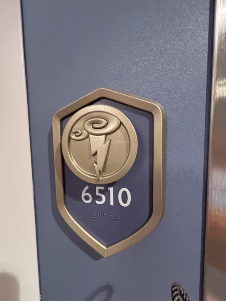 A close-up of the door plaque for Stateroom 6510 on the Disney Destiny, featuring a gold emblem with a lightning bolt and the stateroom number displayed in both standard and braille.