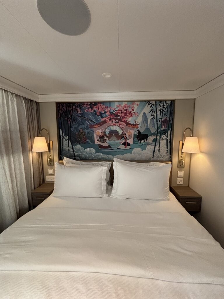 Interior of a Deluxe Family Oceanview Stateroom on the Disney Destiny featuring a large bed with white linens and decorative pillows, flanked by two lamps, and a colorful mural depicting a scenic landscape behind the bed.