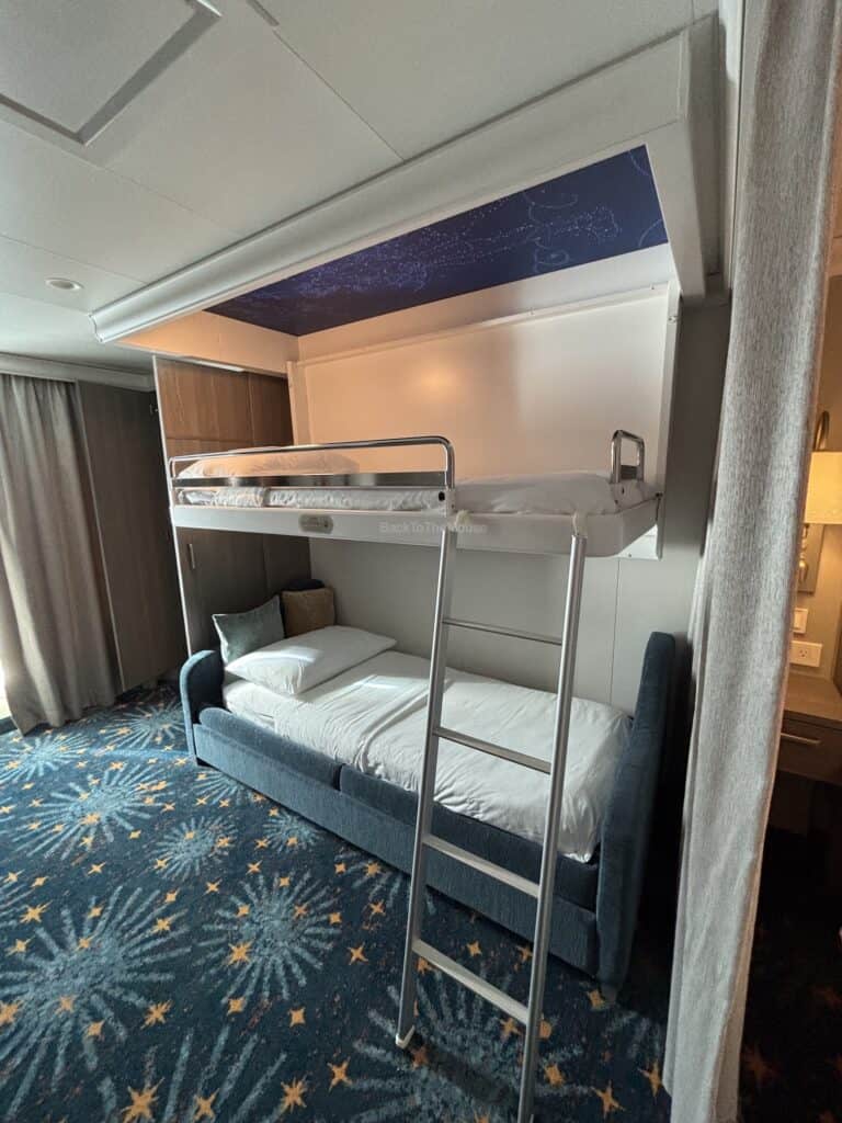 Interior of a Disney cruise ship stateroom featuring a pull-down bunk bed above a sofa bed, with decorative carpet and soft cushions.