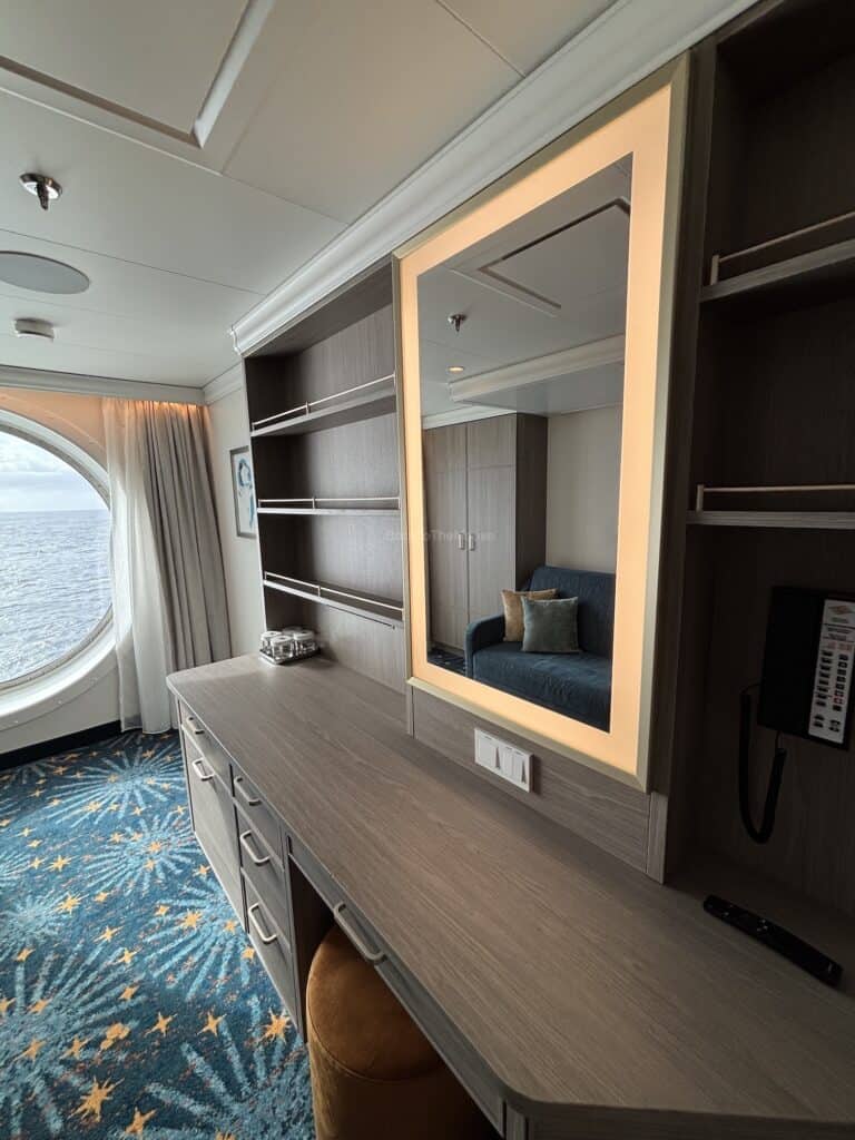 Interior view of a Deluxe Family Oceanview Stateroom on the Disney Destiny, showcasing a desk area with a mirror, a blue couch, and a circular window with ocean views.