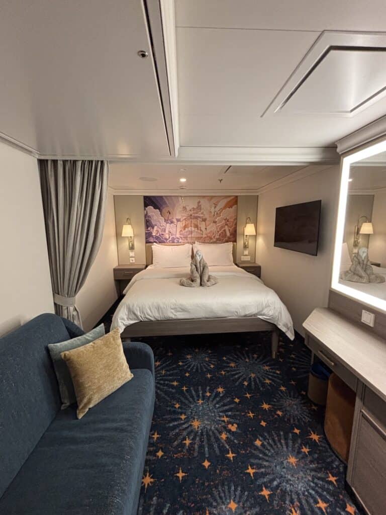 Interior view of a Disney cruise stateroom featuring a bed with decorative towel animals, a blue couch, and a television, complemented by soothing lighting and a patterned carpet.