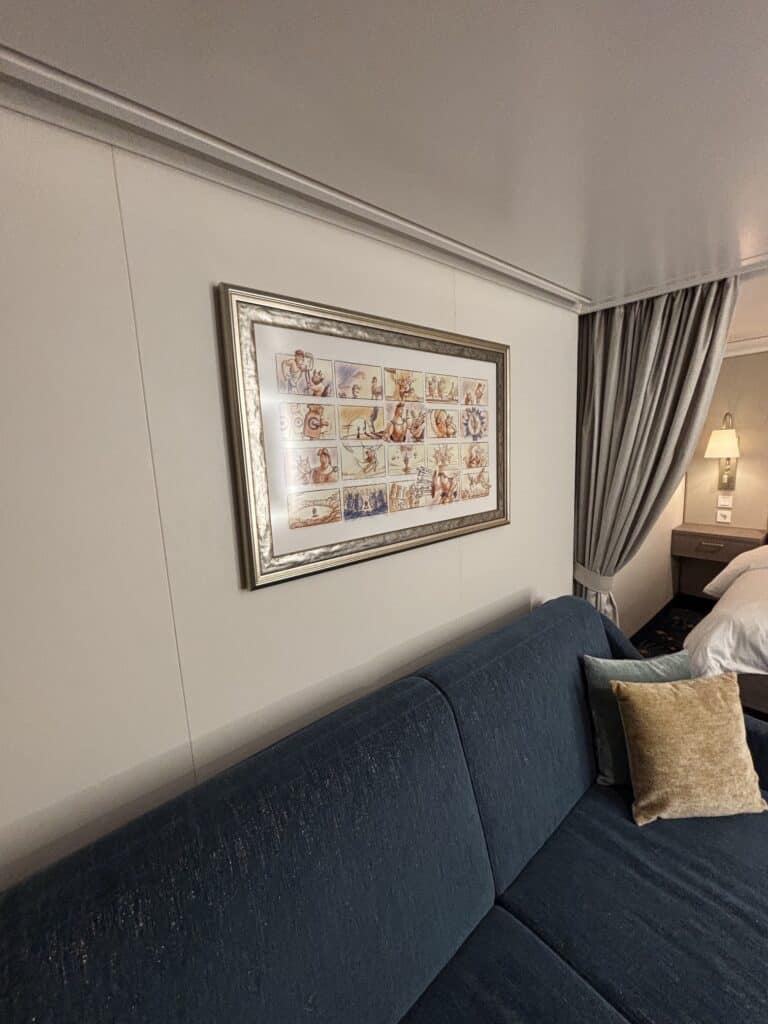 A framed artwork depicting various illustrated scenes hangs above a blue couch in a cruise ship stateroom.