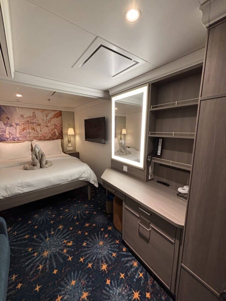 Interior view of a standard inside stateroom on the Disney Destiny, featuring a bed with a towel animal, a nightstand, a television, a desk area, and decorative carpet.
