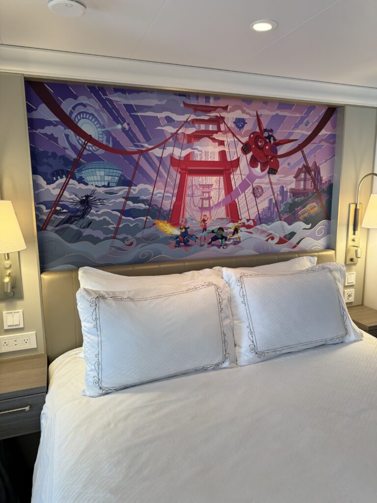 A cozy stateroom on a cruise ship featuring a large bed with decorative pillows, illuminated by bedside lamps, and a vibrant mural depicting characters and scenes from popular cartoons.