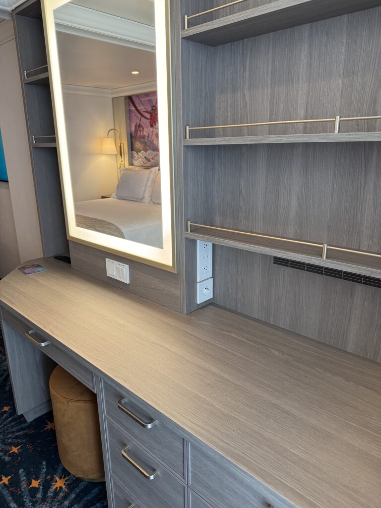 A well-organized dresser area with a lighted mirror, showcasing shelves and electrical outlets, in a Disney Cruise stateroom.