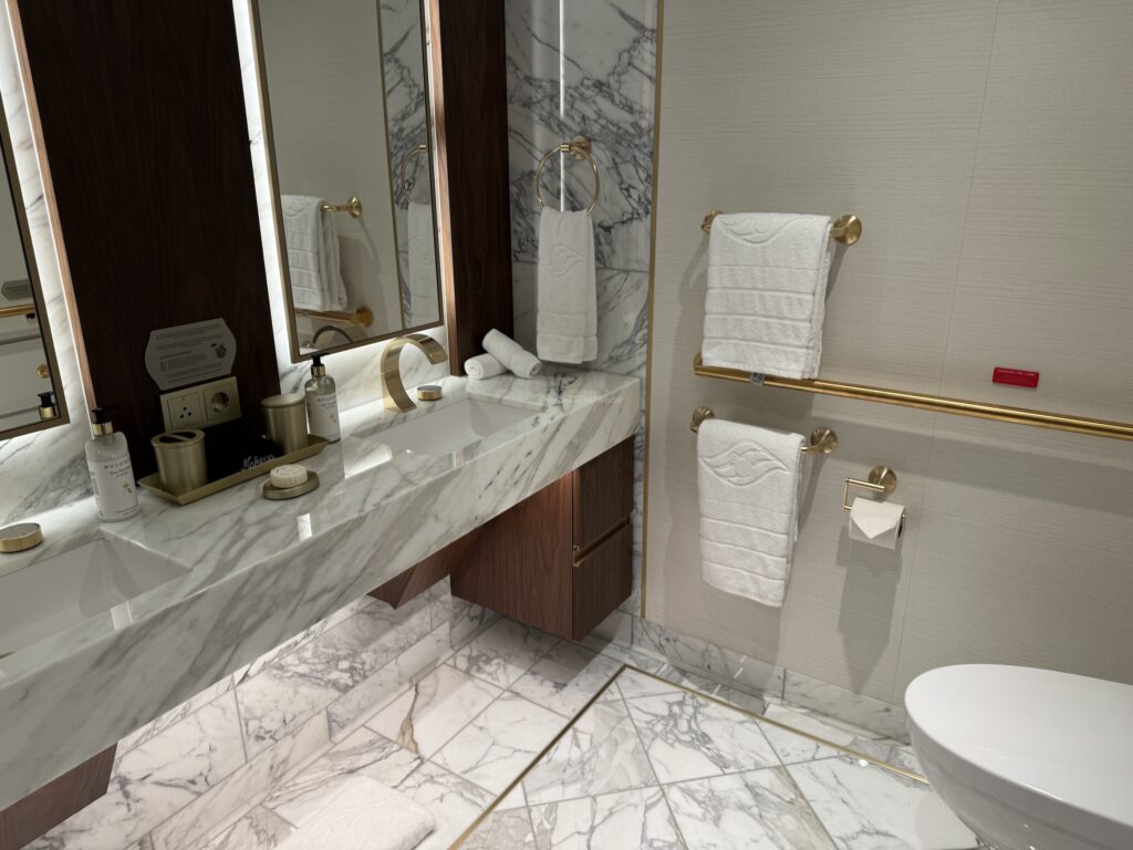 A luxurious bathroom in a cruise suite featuring a marble countertop, dual sinks, vanity mirrors, toiletries, and neatly arranged towels.