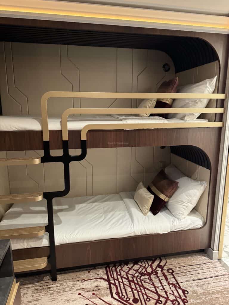 Interior view of a bunk bed setup in a stateroom, featuring a lower and upper sleeping area with decorative pillows, set against a modern backdrop.