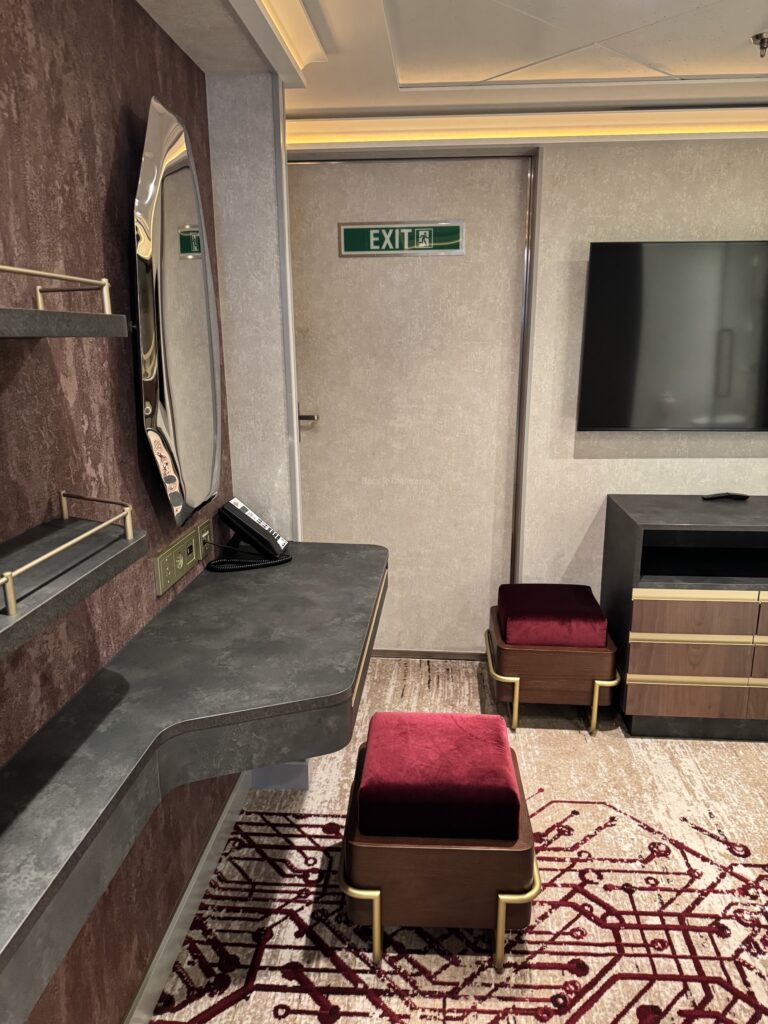 Interior view of a modern cruise ship stateroom featuring a desk area with a mirror, a television, and two red cushioned stools on a patterned carpet.
