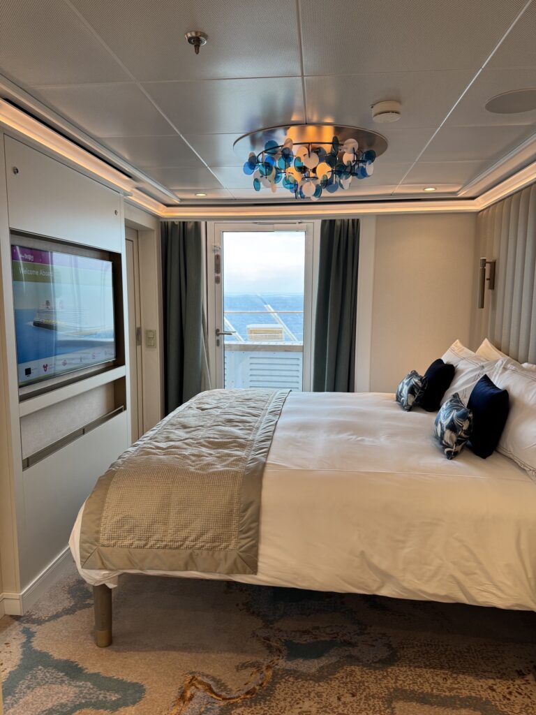 Interior view of a cruise ship stateroom featuring a large bed with decorative pillows, modern lighting, and a window showing the ocean view.