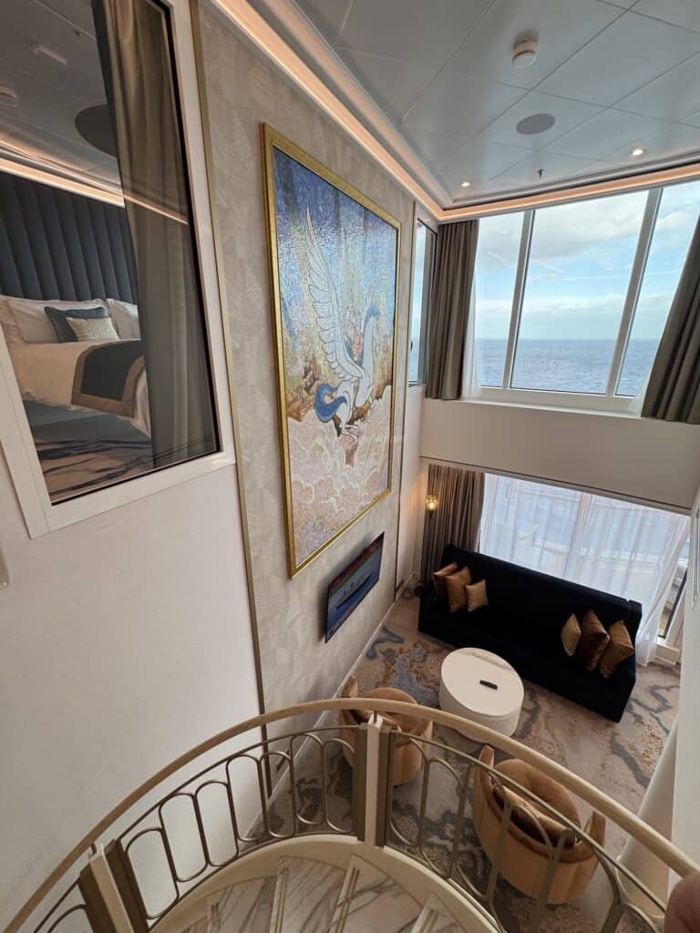 Interior view of a luxurious cruise suite featuring a large wall mural of a winged figure, a spiral staircase, and a sitting area with a black sofa and modern decor, large windows showcasing ocean views.