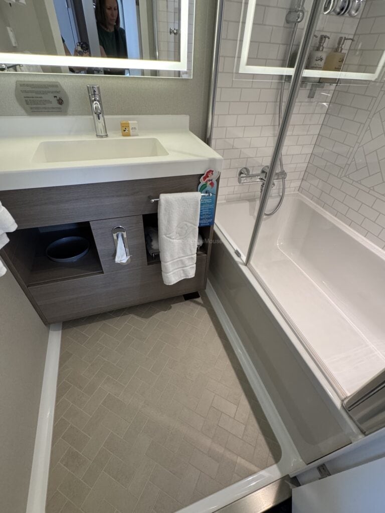Interior view of a bathroom in a Deluxe Oceanview Stateroom on Disney Destiny, showing a sink area with a mirror, storage cabinet, and bathtub with shower.