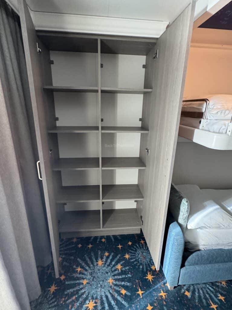 Interior view of an open closet with shelves in a stateroom on the Disney Destiny, showing the carpet and a couch nearby.