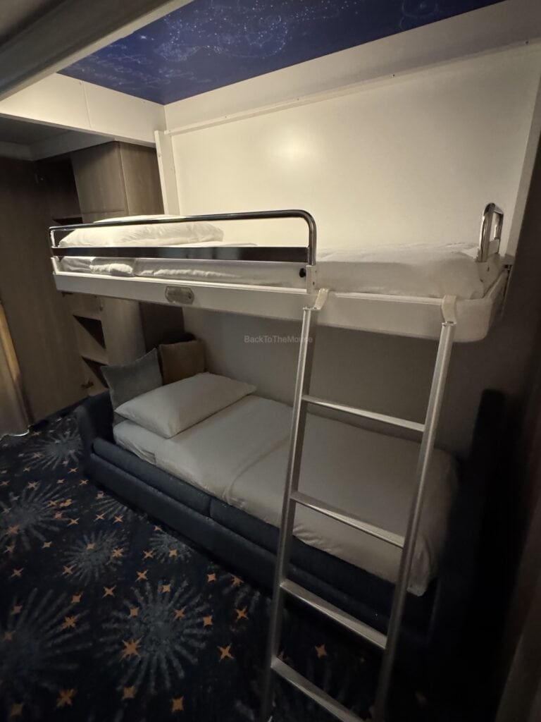Interior view of an 8C Deluxe Family Oceanview Stateroom on the Disney Destiny, featuring a lower single bed and an upper pull-down bed, with a starry carpet design.