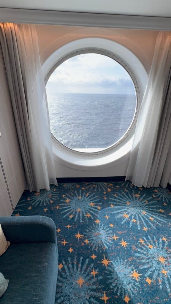 Interior view of a Deluxe Family Oceanview Stateroom on the Disney Destiny, featuring a round window with ocean views, light curtains, and a patterned carpet.