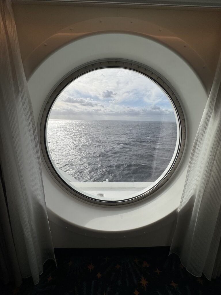 View of the ocean through a circular window in a cruise ship stateroom, with partly cloudy sky visible.