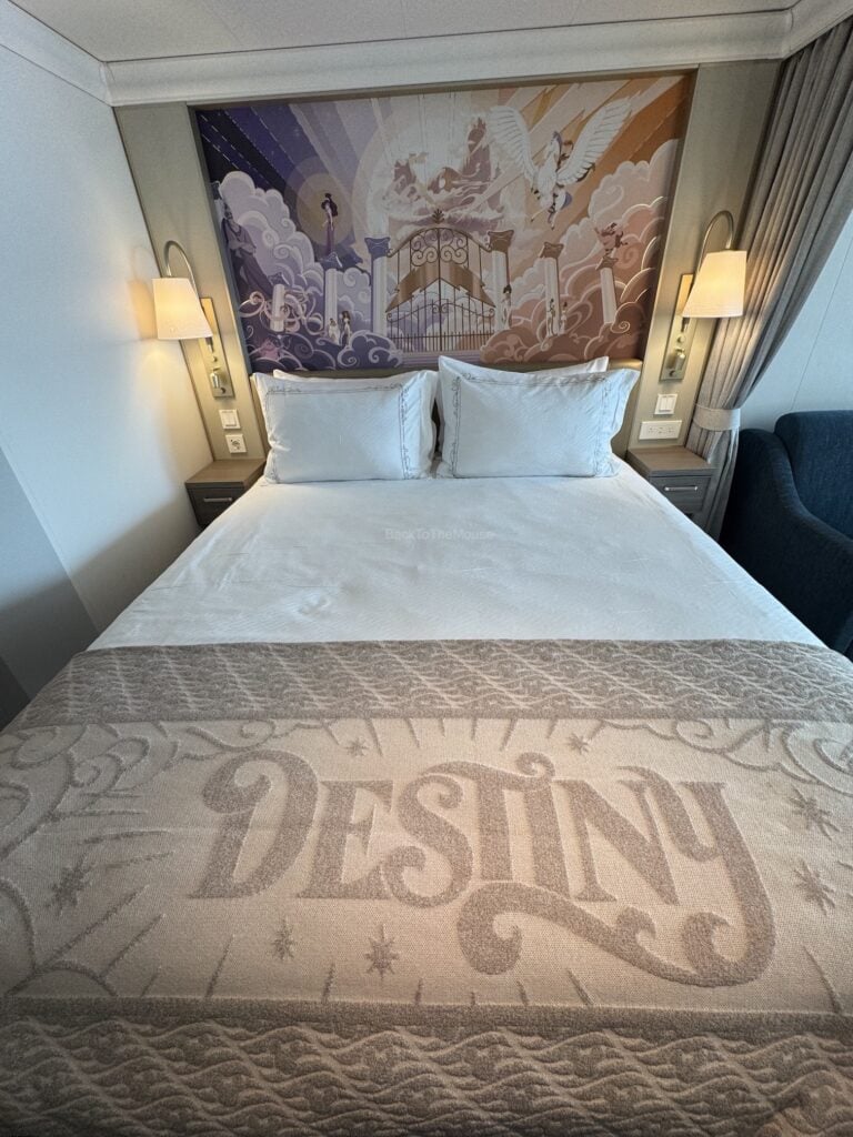 A view of a stateroom on the Disney cruise ship, featuring a large bed with a decorative blanket that reads 'DESTINY' and a colorful mural of clouds and characters on the wall behind the bed.