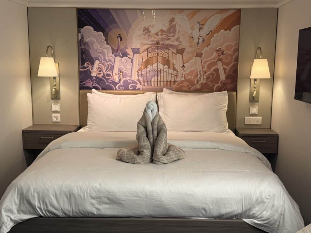 Cozy interior of a Disney cruise stateroom with a neatly made bed, soft gray bedspread, and a decorative towel sculpture on top. The wall features a colorful mural depicting whimsical scenes, and there are bedside lamps and modern fixtures.
