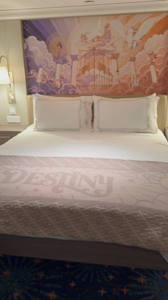 Interior view of a Disney cruise stateroom featuring a bed with decorative pillows, a patterned blanket, and a colorful mural in the background.