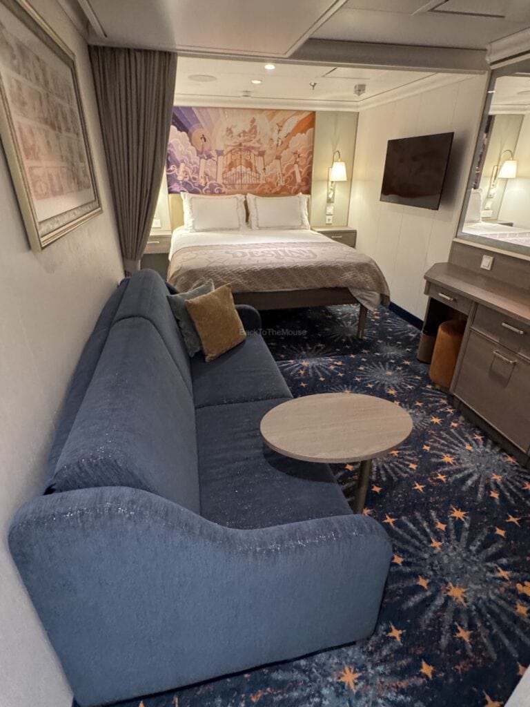 Interior view of a standard inside stateroom on the Disney Destiny, featuring a queen bed, a blue couch, a round coffee table, and a large television.